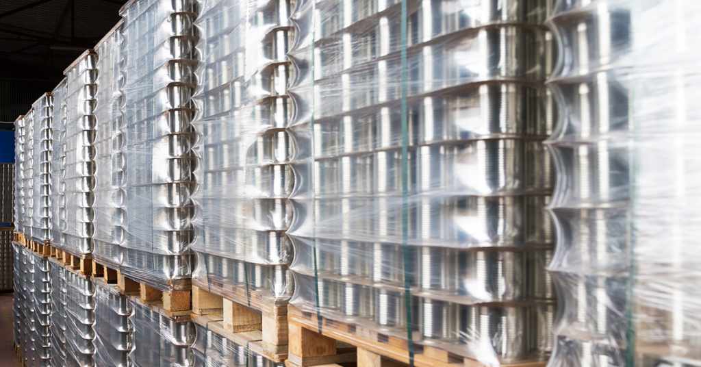 Highly stacked wooden pallets in a warehouse, loaded with hundreds of silver aluminum cans. Each pallet is tightly wrapped in clear protective plastic film, creating a shimmering, reflective surface. The stacks extend deep into the background, illustrating a large-scale beverage supply chain or manufacturing facility.