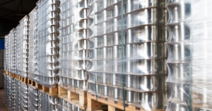 Highly stacked wooden pallets in a warehouse, loaded with hundreds of silver aluminum cans. Each pallet is tightly wrapped in clear protective plastic film, creating a shimmering, reflective surface. The stacks extend deep into the background, illustrating a large-scale beverage supply chain or manufacturing facility.