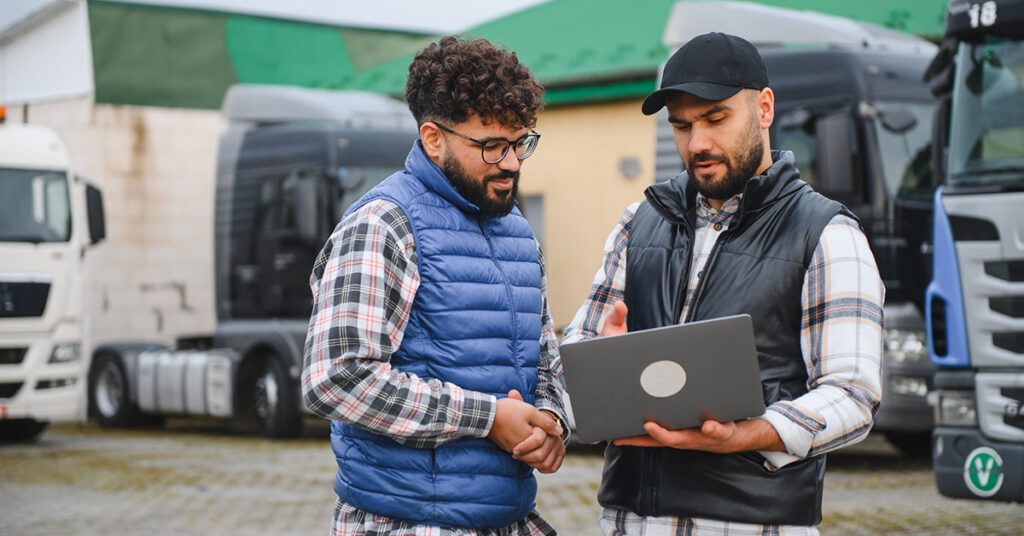 two people having a discussion and looking at computer next to a few semi trucks