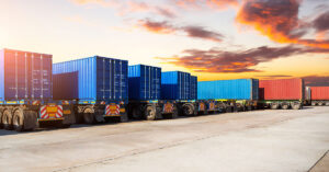 A row of shipping containers loaded on semi-truck trailers parked in a lot under a sunrise or sunset sky.