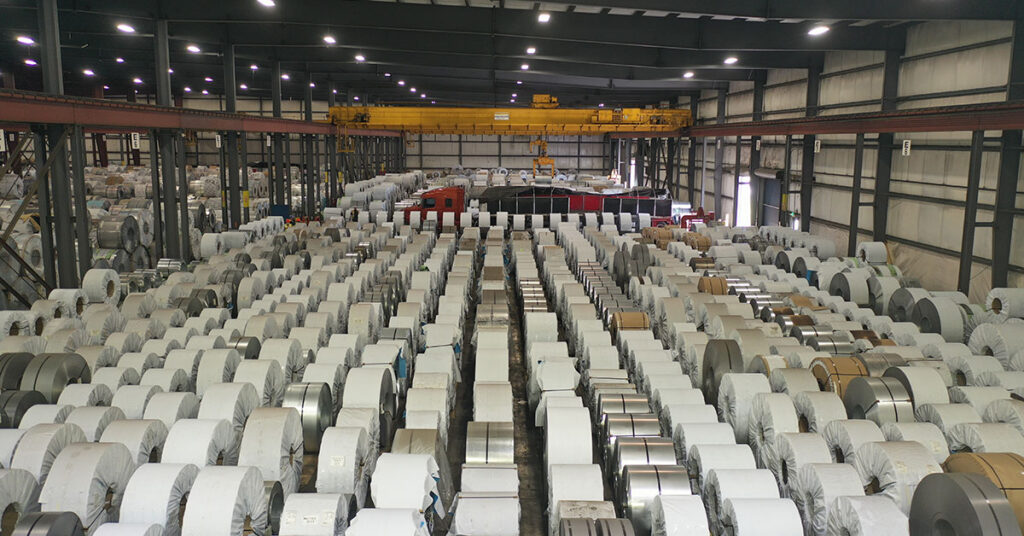 A warehouse interior featuring rows of large, metallic industrial coils stored in racks, with a yellow overhead crane visible above.