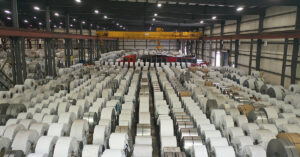 A warehouse interior featuring rows of large, metallic industrial coils stored in racks, with a yellow overhead crane visible above.