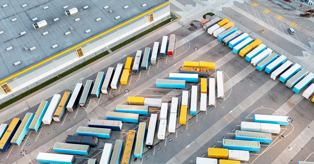 Aerial high-angle view of a large logistics shipping yard with dozens of semi-truck trailers in blue, yellow, and white organized in neat diagonal parking rows next to a large industrial warehouse.