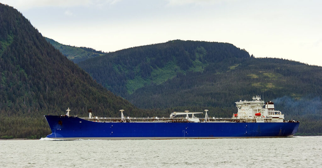 A blue industrial freighter vessel sailing past forested coastal mountains in Alaska.
