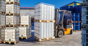 Forklift moving stacked IBC tote tanks in an industrial yard.