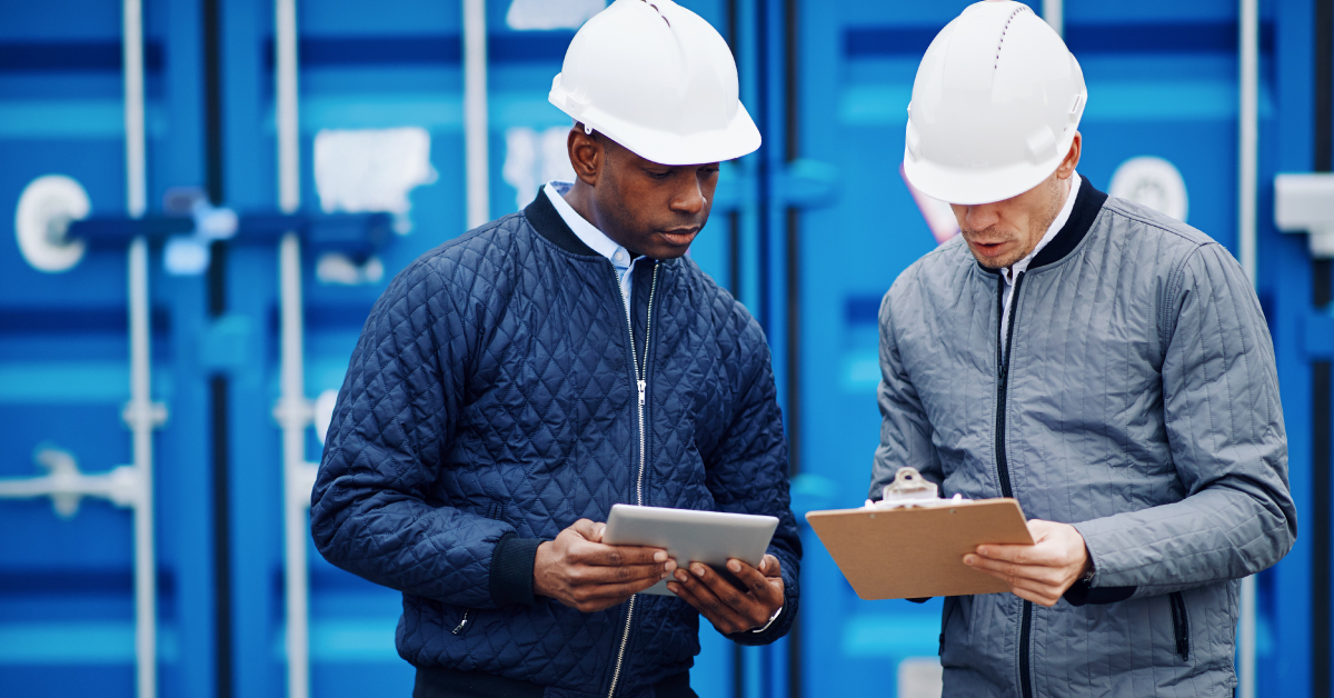 two men standing in front of a cargo shipping container wearing hard hats and looks at clipboards in their hands