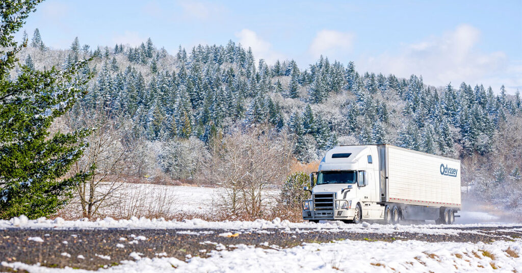 Big rig long haul white semi truck tractor transporting commercial cargo in refrigerator semi trailer going on the wet glossy road with water from melting snow and winter snowy trees on the hill