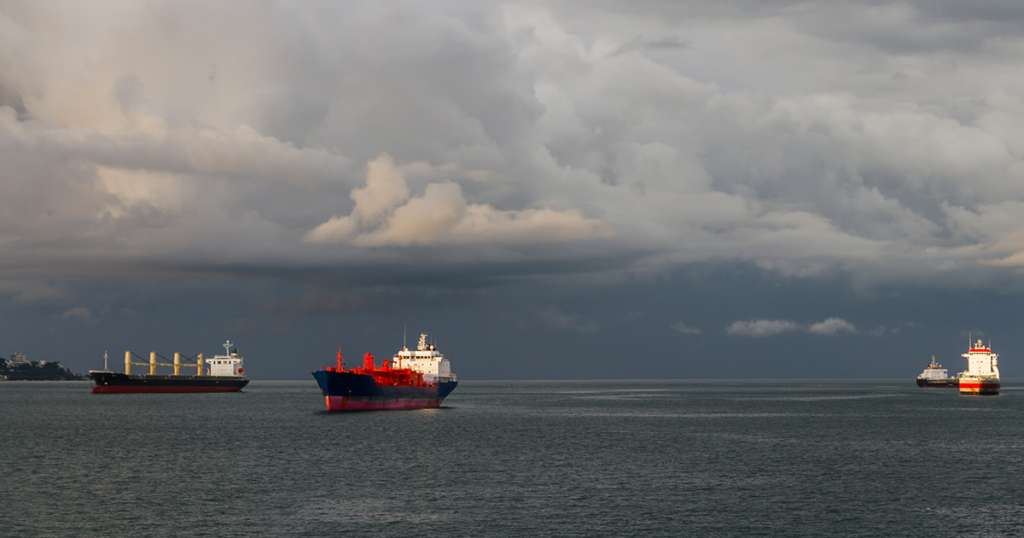 Four cargo ships stationed in the water as the sky is filled with dark storm clouds.